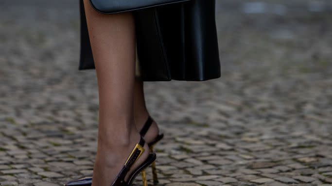 a woman wearing sheer black tights with pointed toe slingback heels, a black leather shoulder bag, and a long black coat on a cobblestone street in berlin