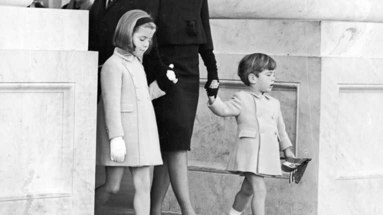 jacqueline kennedy holds the hands of her children caroline and john f kennedy jr as they walk down steps, jacqueline wears all black with a veil and the children wear light colored peacoats