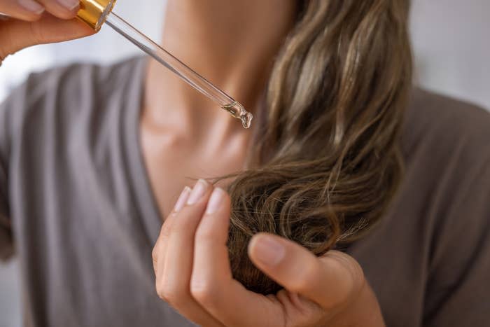 A person applies oil to the tips of their hair using a dropper. The person wears a casual shirt