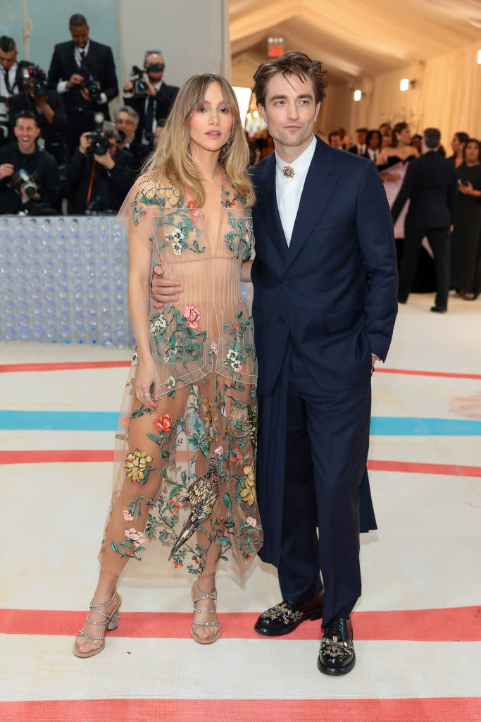 Two celebrities on red carpet; one in a sheer floral dress, the other in a navy suit with embellished shoes