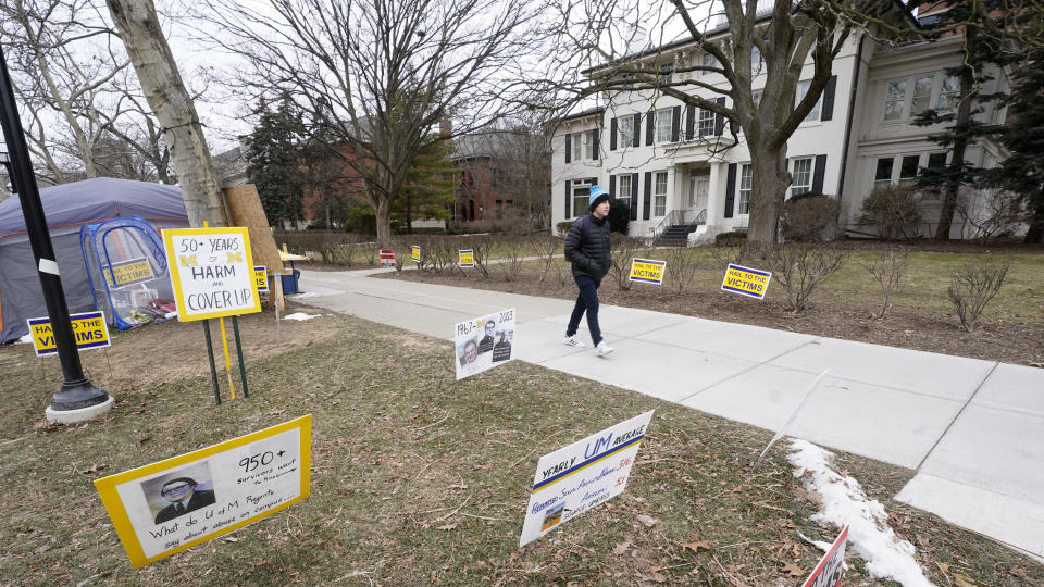 A student walks past signs outside the University of Michigan's Presidents House on campus in Ann Arbor, Mich., Thursday, Jan. 20, 2022. A financial payout for more than 1,000 people — mostly men — who say they were sexually assaulted by former University of Michigan sports doctor Robert Anderson is the latest multimillion-dollar settlement involving schools faced with sexual misconduct scandals. (AP Photo/Paul Sancya)