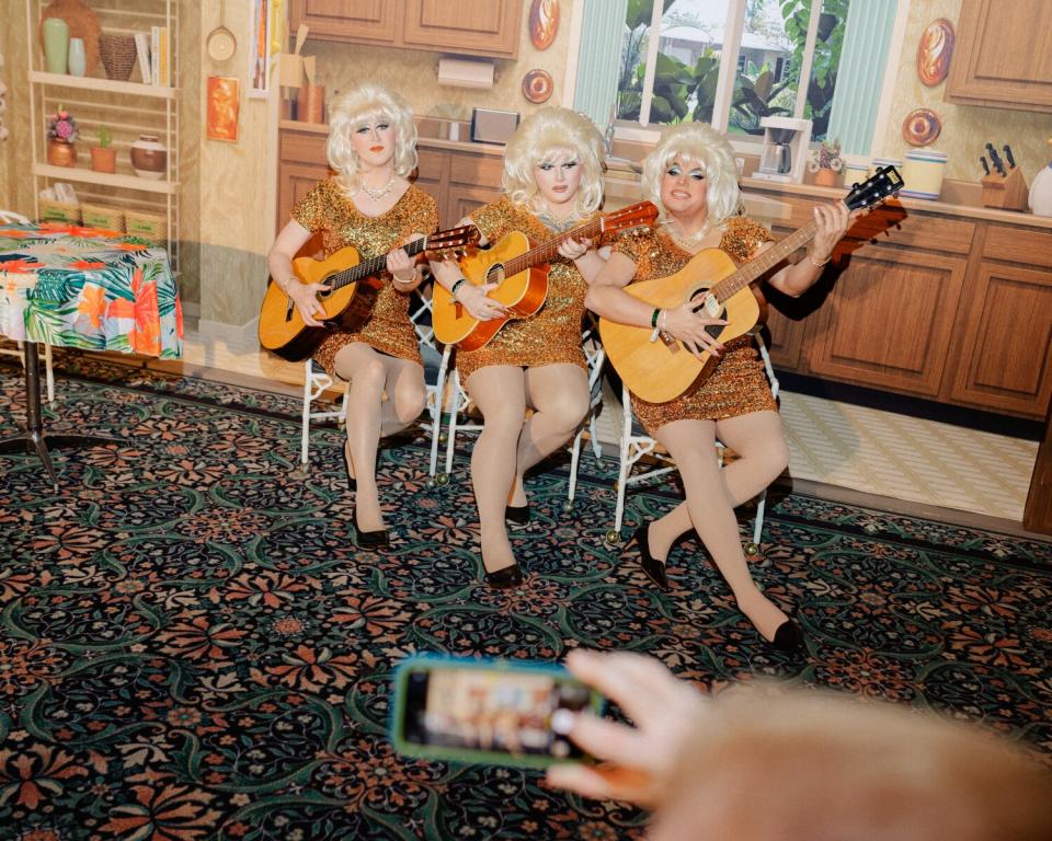 Three people in drag play guitar in front of a kitchen backdrop.