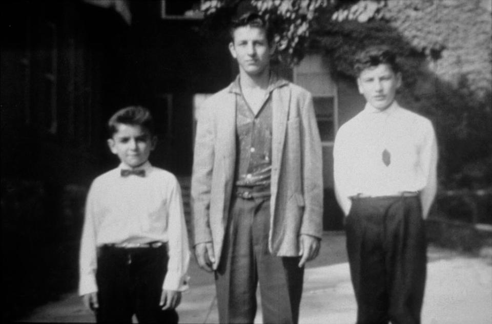 Three young boys stand outdoors in front of a building. The boy on the left wears a bow tie, the boy in the middle wears a jacket, and the boy on the right wears a tie