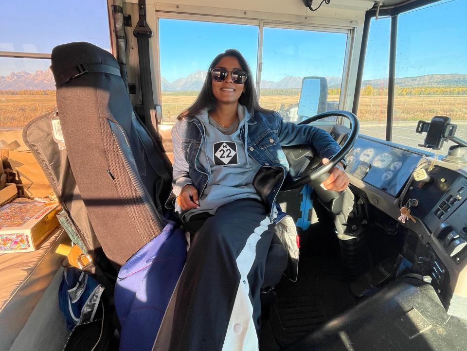 A woman sits in front driver's eat of school bus and smiles with blue sky and desert area in background