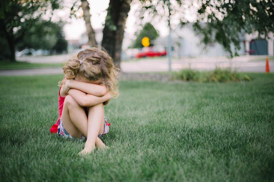 Little girl with curly hair sits on grass and cries into her arms.