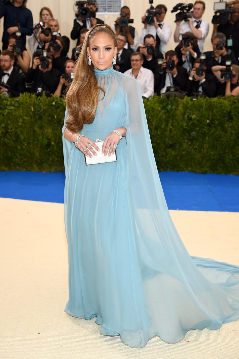 Jennifer Lopez in a flowing blue gown with sheer overlay and high neckline, at a gala event with photographers in the background