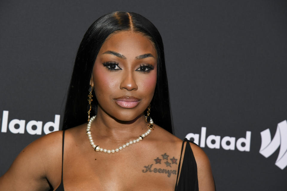 Kash Doll attends a GLAAD event, wearing a black dress with a pearl necklace and long, straight hair. She has star tattoos on her chest