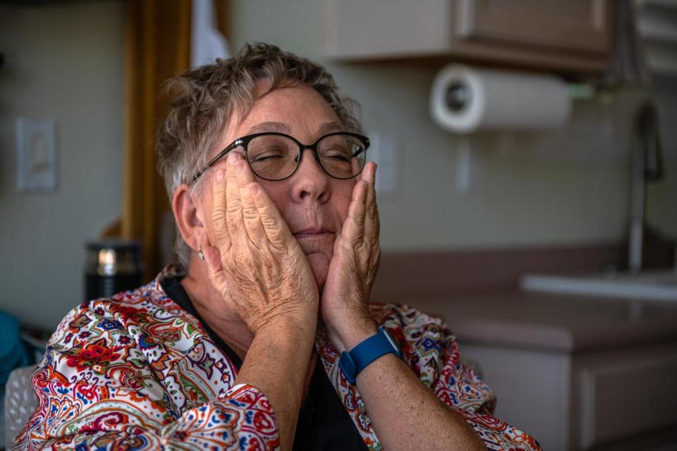 A woman with short hair and glasses, wearing a flowery shirt, holds her hands up to her cheeks