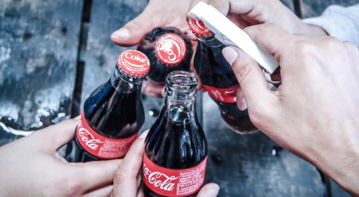 Close-up photo of hands holding glass Coca Cola (KO) bottles, clinking them together. One hand has a bottle opener and is opening a bottle.