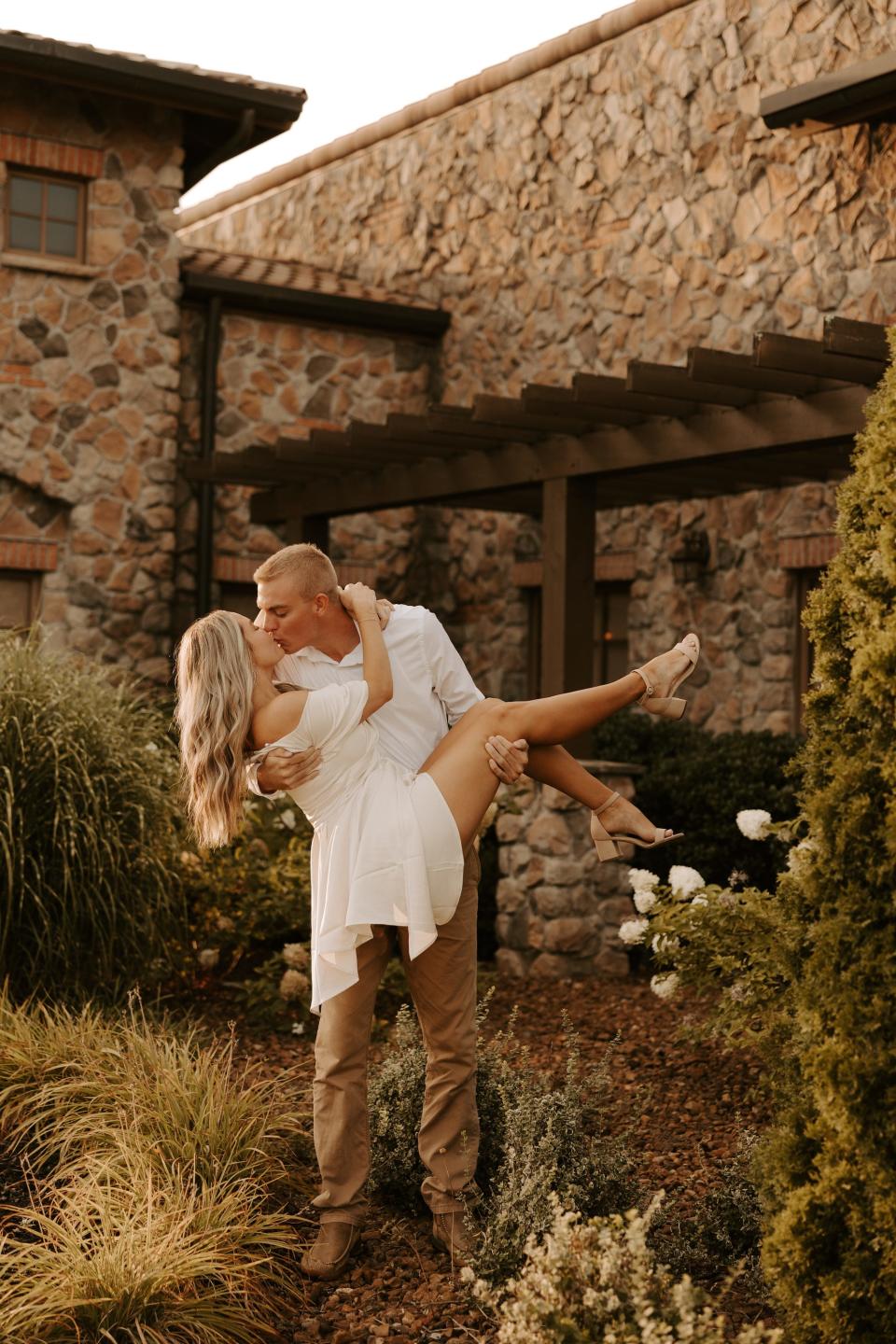 A man in a white shirt and khakis carries and hugs a woman in a white dress in front of a stone building.