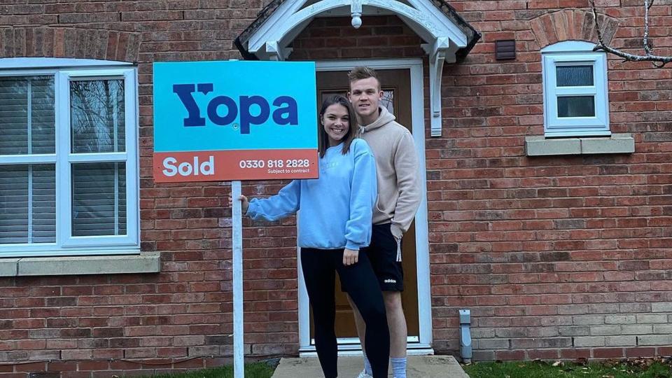 Chloe Hewitt and George Betts holding sold sign outside house