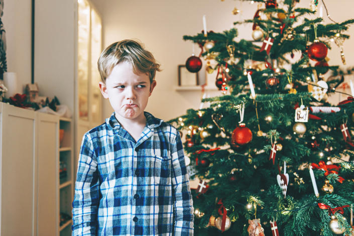A sad boy wearing pajamas standing in front of a Christmas tree.