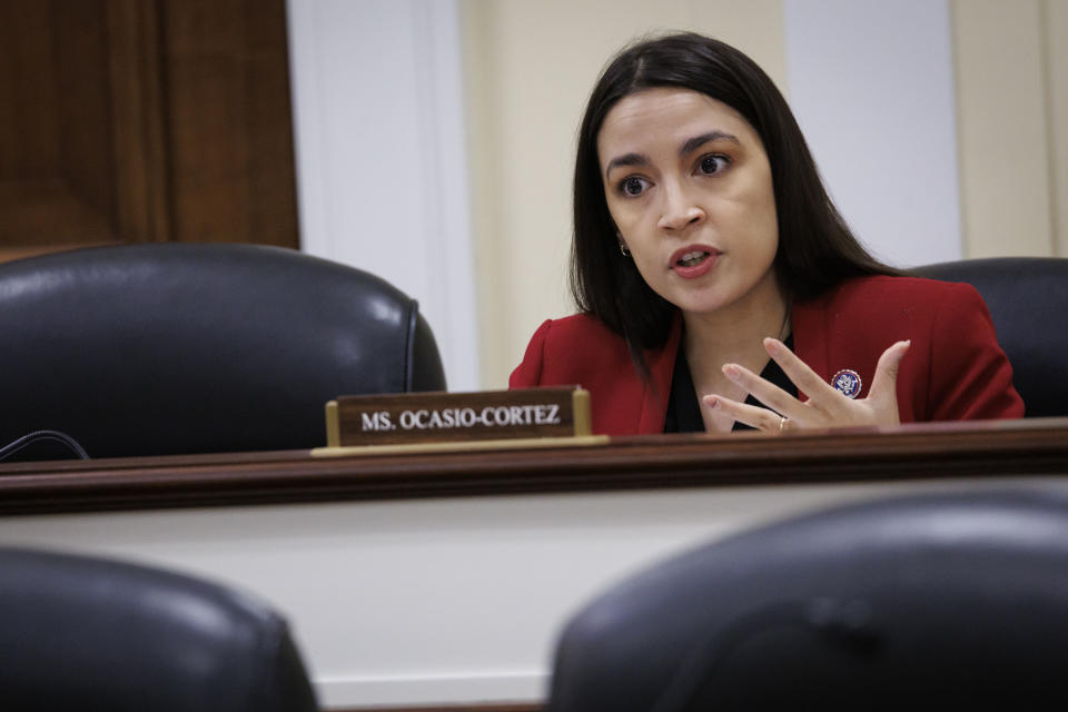 Rep. Alexandria Ocasio-Cortez sits at a long desk behind a placard bearing her name.