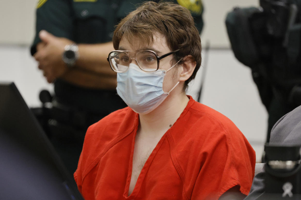 Nikolas Cruz, in orange jumpsuit, and large glasses above a surgical mask, with officers standing behind him, shows no emotion. 