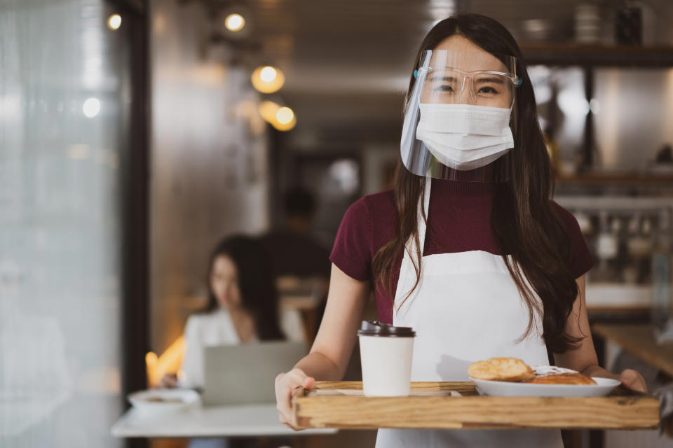 Asian waitress with face protective mask and face shield serving coffee and bread in coffee shop