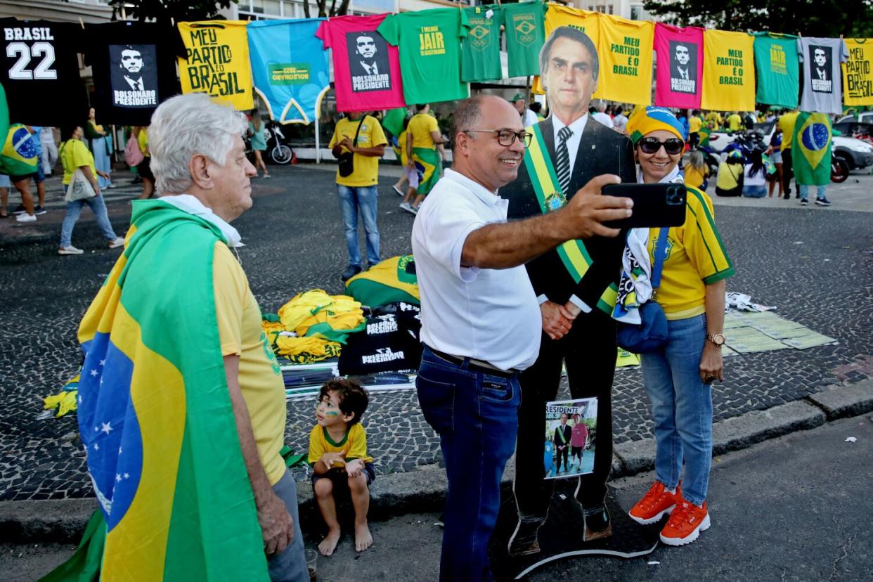 A man takes a cellphone photo of a woman standing next to a cardboard image of a man in a suit with a green-and-yellow sash