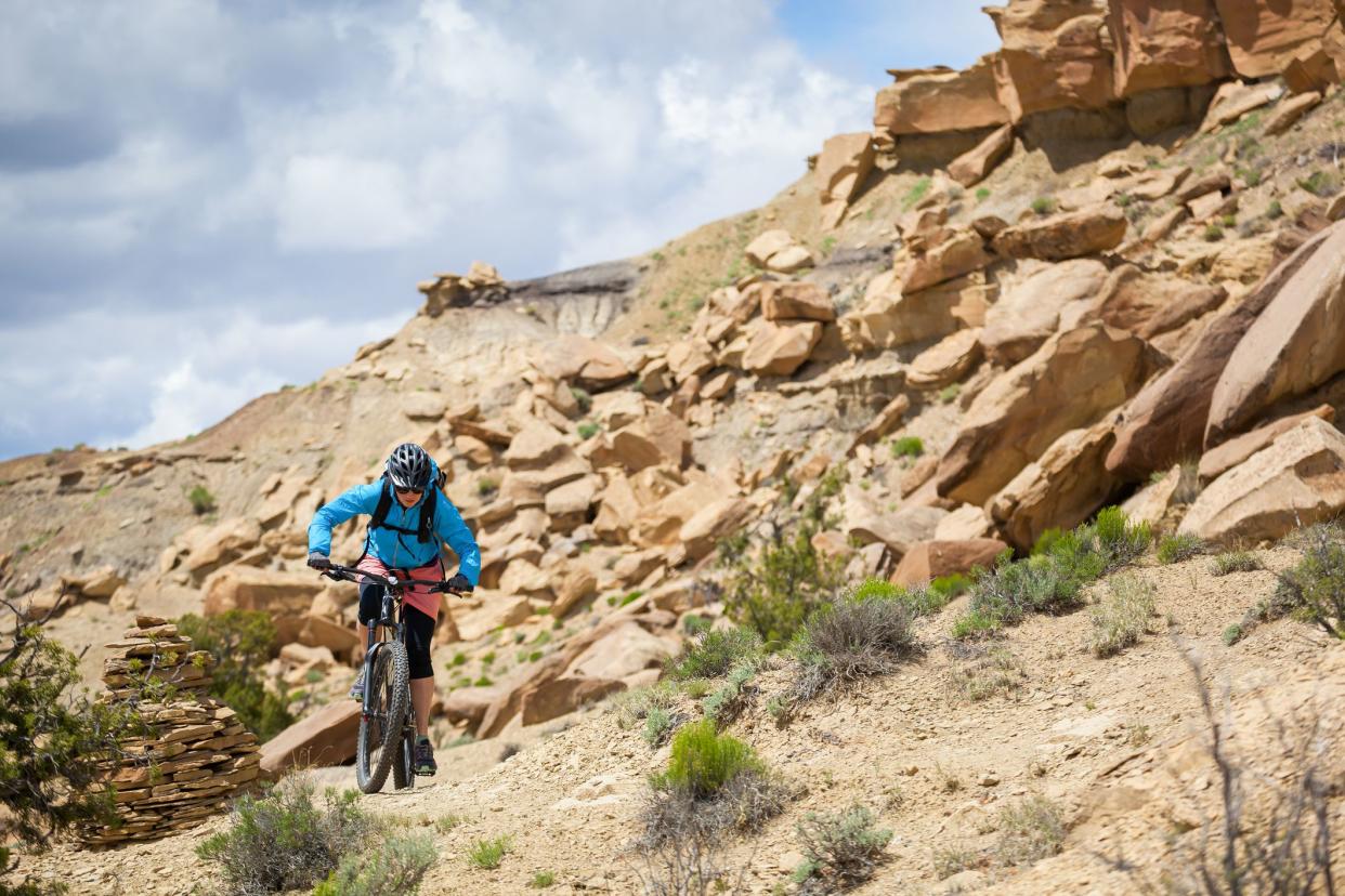 a woman pedals in the deserts badlands landscape underneath a cloud filled blue sky. horizontal composition taken in gallup, new mexico.