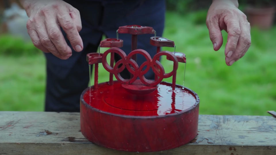 The YouTuber, "Grandpa Amu," standing behind a red, wooden water fountain with a levitating Olympics symbol.