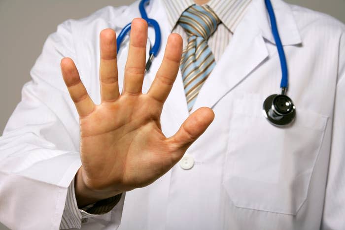 A doctor wearing a lab coat and stethoscope holds up his open hand towards the camera, signaling to stop. His face is not shown