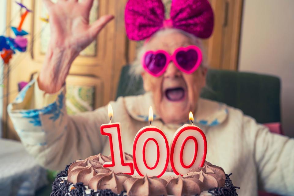 An older woman in novelty glasses behind a cake with candles that say "100."