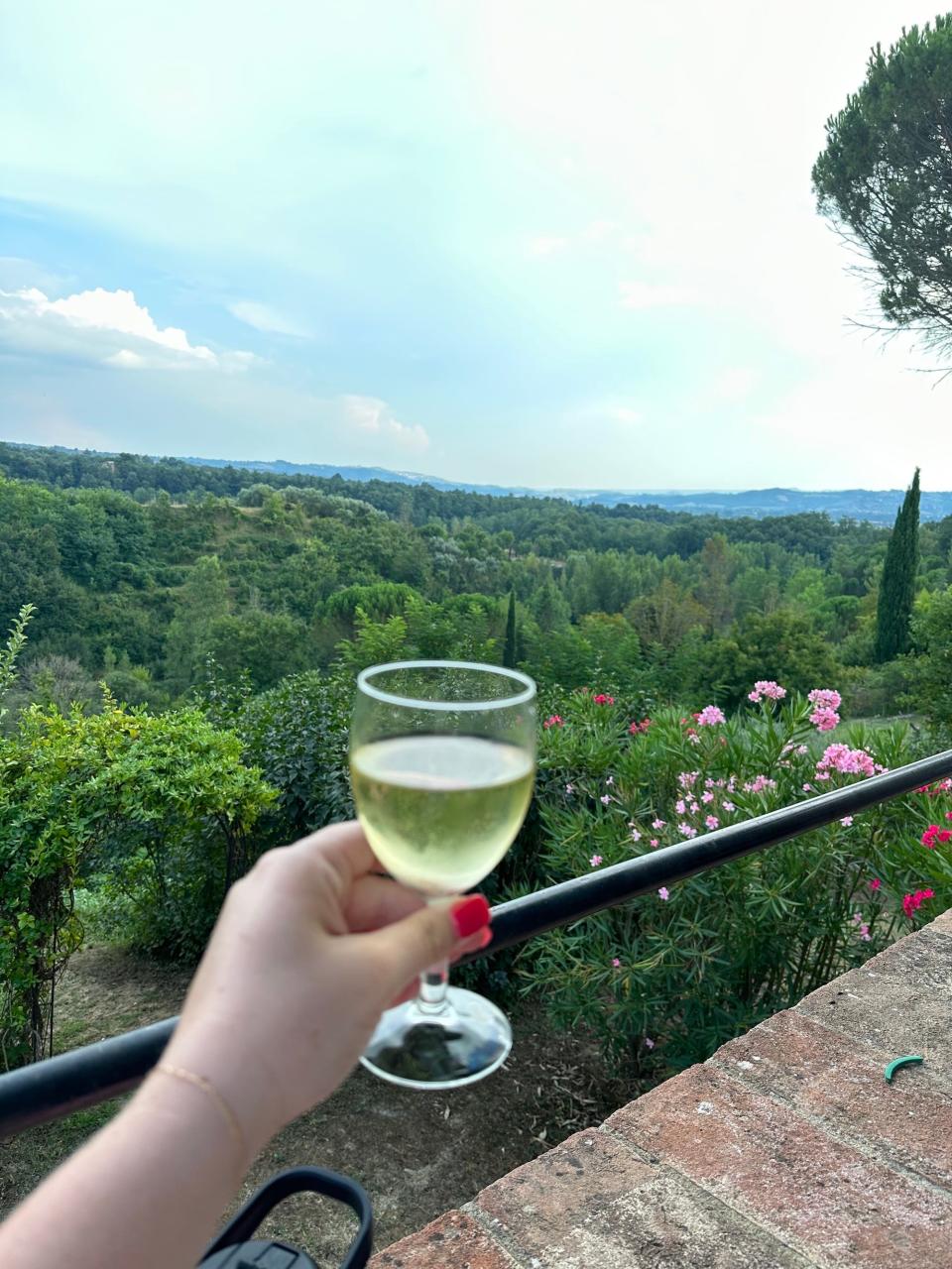 A woman holds up a glass of wine from a porch on an Italian villa.