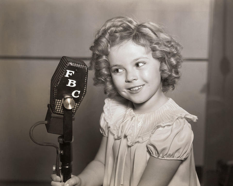 Shirley Temple, a young girl with curly hair, smiles while standing at a vintage microphone marked "FBC." She is wearing a dress with a lacy collar