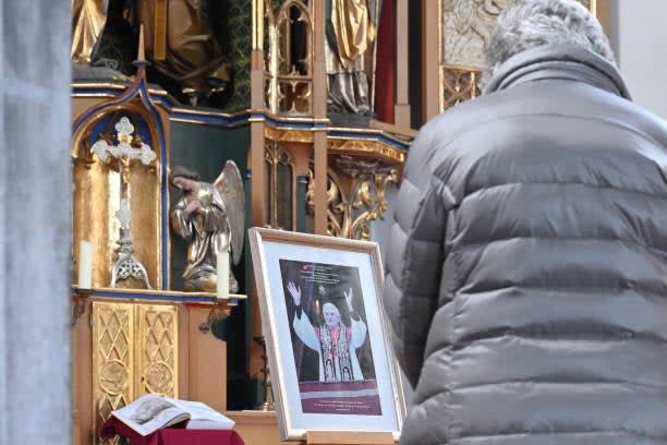 A believer stands in front of a picture of Pope Emeritus Benedict XVI set up next to a prayer bench at the Catholic St Oswald church in Marktl, southern Germany, on 29 December 2022 (AFP via Getty Images)