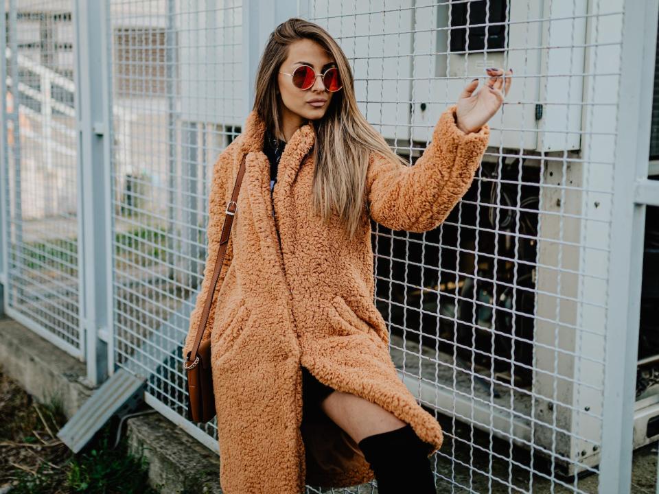 A woman in a faux fur coat stands in front of a fence.