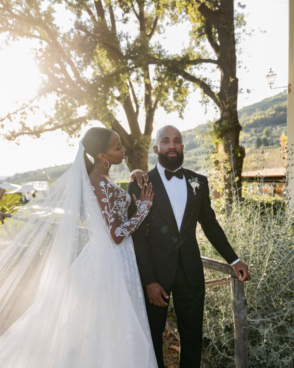 A bride leans her hand on her groom and looks at him. They stand in front of greenery.