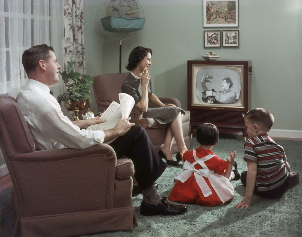 A happy family cheerfully sits in their living room and watches a televisied clown and puppet show, 1957. The father holds an newspaper open to the financial section in his hands.