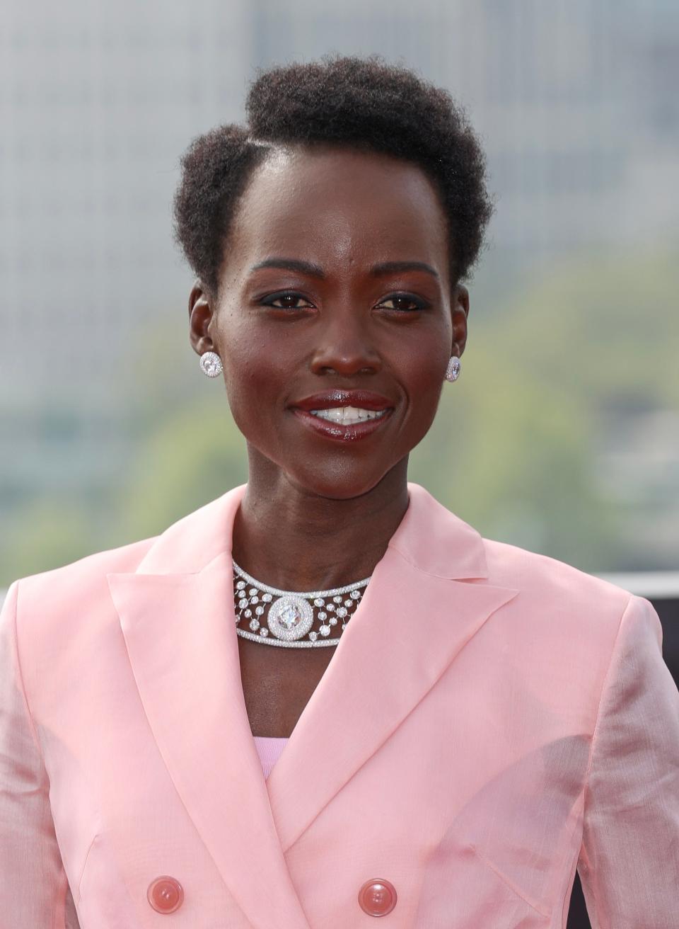 Lupita Nyong'o wearing a chic tailored suit jacket and a statement necklace, photographed at an outdoor event