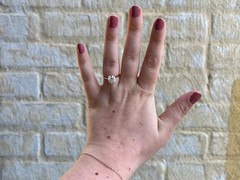 A hand with an engagement ring on it sits in front of a brick wall.