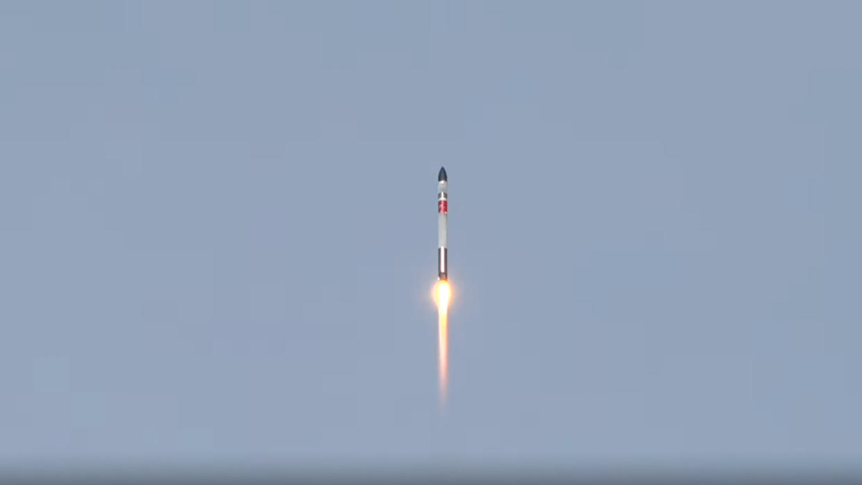  A white, black and red rocket lab electron rocket launches into a blue sky. 