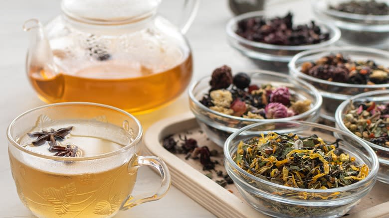 Glass cup and teapot full of tea next to dishes of dried herbal blends