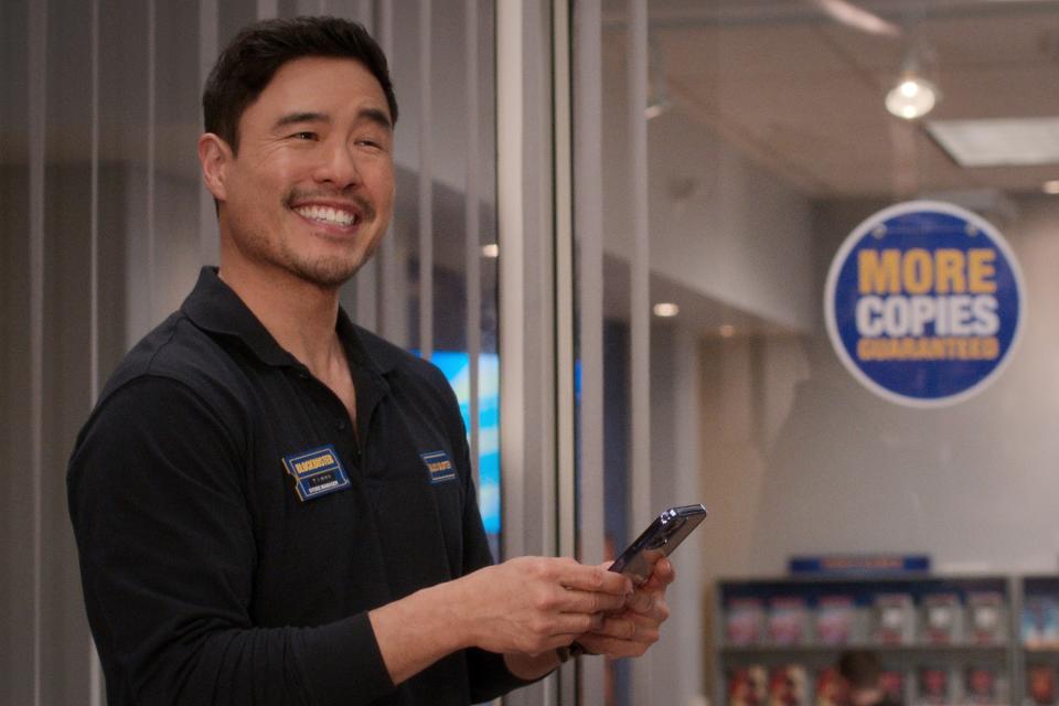 Randall Park in a scene in a store uniform smiling at his phone with a "More Copies" sign in the background