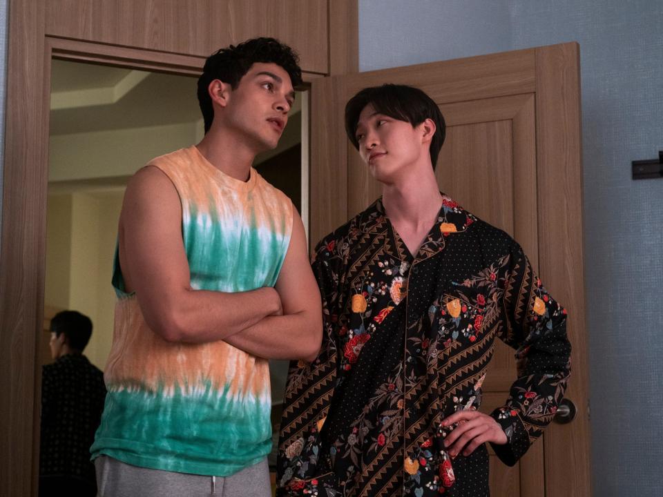 Q, played by anthony keyvan, and min ho, played by sang heon lee, in xo kitty. Q is wearing a tie-dyed tank and crossing his arms, while minho is in a patterned button up shirt and looking up at Q