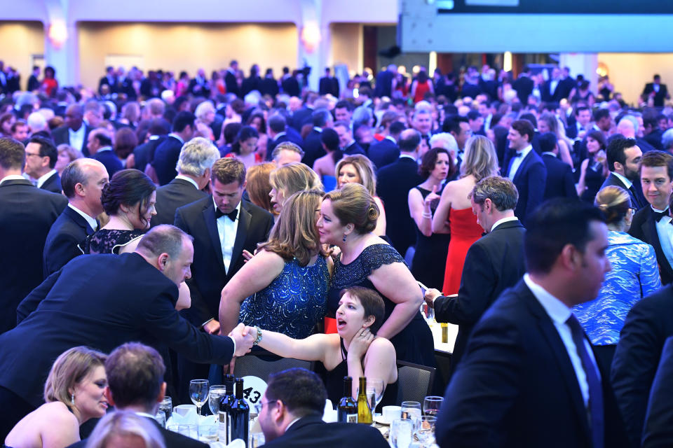 Hundreds of people in formal attire are seated or standing while mingling in a large room.