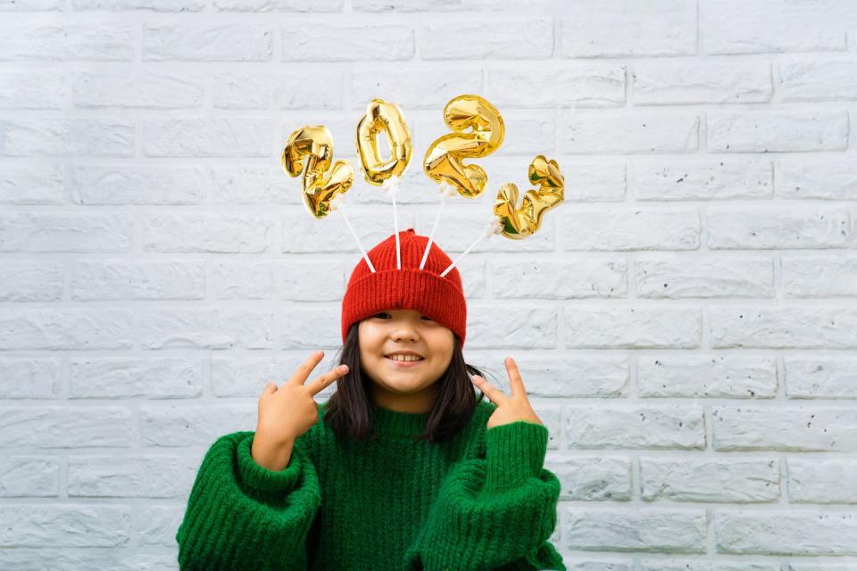 happy asian baby girl in a green sweater close up with golden balloons with numbers 2023 in a hat copy space, white brick wall background, girl looking at the camera, smiling