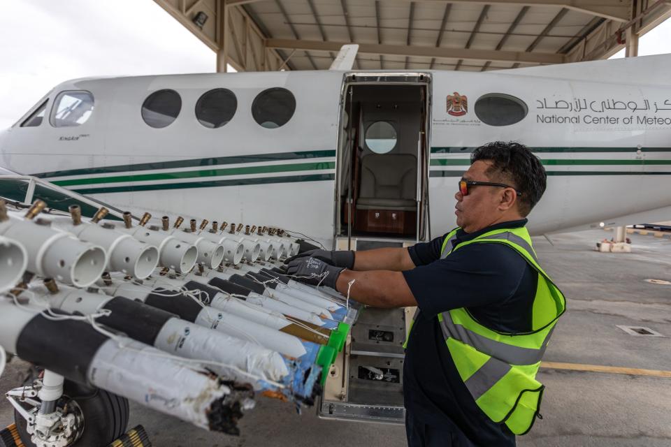 man in black shirt neon green vest handles a row of canisters in a mounting device on the wind of a small white plane