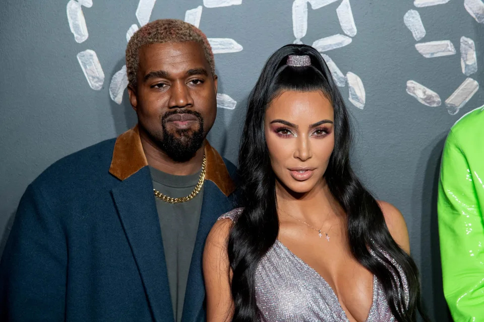 Kanye West and Kim Kardashian at an event; Kanye is in a coat and chain necklace, and Kim is in a glamorous dress with a high ponytail