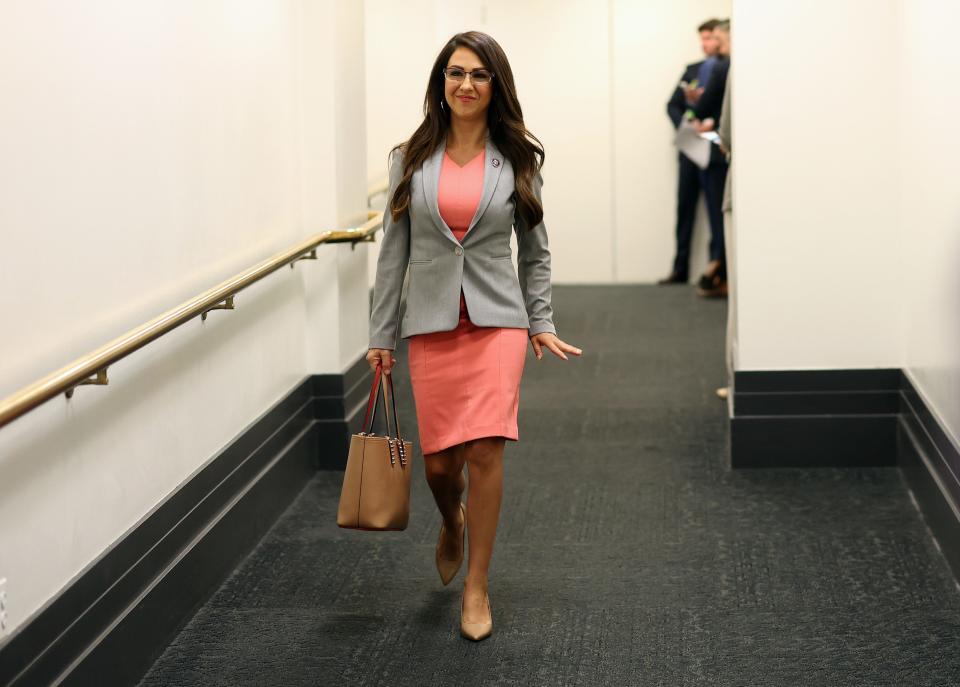 Lauren Boebert in a pink dress, black glasses, a grey blazer and brown bag walks down a white-walled hallway on gray carpeting.