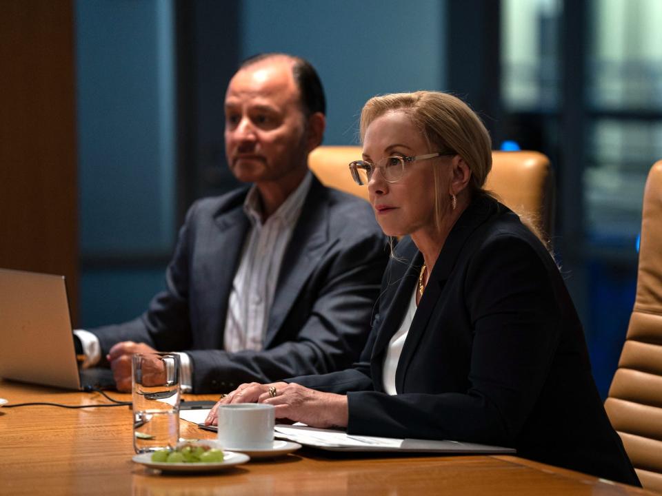 A middle aged white man and white woman wearing navy suits sitting at a brown conference table in an office.