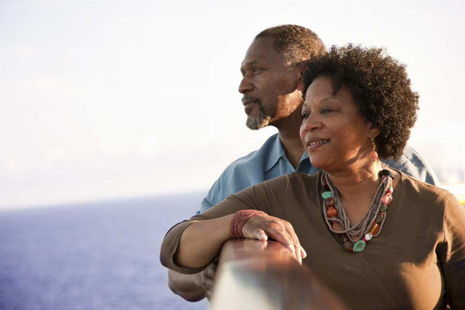 An older couple, casually dressed, smiles while leaning on a railing and looking at the ocean, enjoying a peaceful moment together