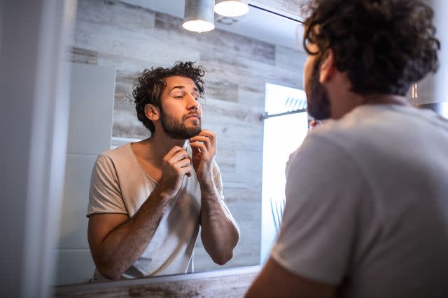 <p>stefanamer / Getty Images</p> Male with beard looking at mirror and touching face in bathroom