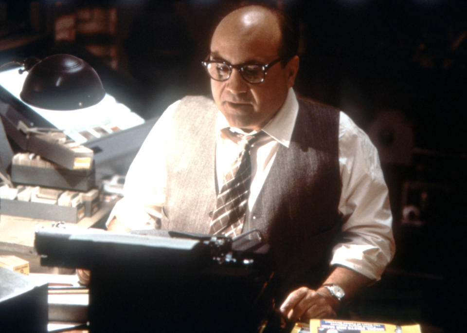 A balding man with glasses and wearing a vest and tie types on a typewriter in a dimly lit office. His desk is cluttered with papers and books