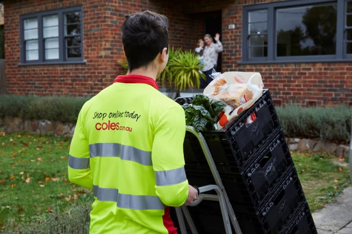 A Coles delivery driver arrives at a woman's home as she waves to him. He's pushing crates.