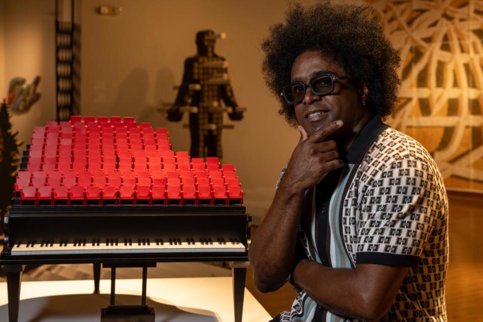 A man wearing a geometric shirt stands in front of a sculpture in the shape of a piano in a gallery