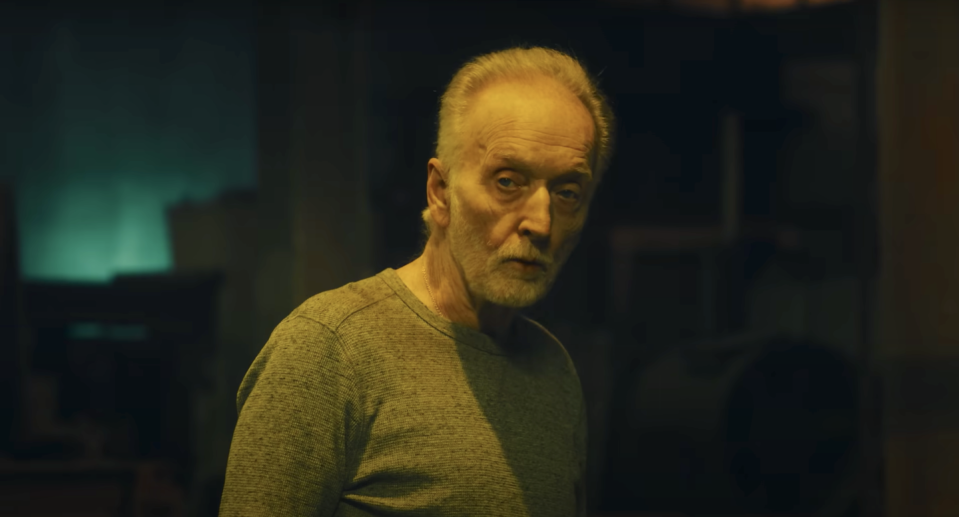 tobin bell as jigsaw in saw x, an older man with grey hair and beard stands in a dark room looking at the camera, he wears a long sleeve grey top