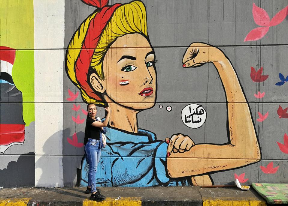 In this Monday, Nov. 18, 2019 photo, a woman poses for a photo in front of graffiti at the Saadoun tunnel, in Baghdad, Iraq. The tunnel that passes under Baghdad’s landmark Tahrir Square has become an ad hoc museum for Iraq's revolution: Young artists draw images and murals that illustrate the country’s tortured past, and the Iraq they aspire to. Arabic reads, "those are our women." (AP Photo/Khalid Mohammed)