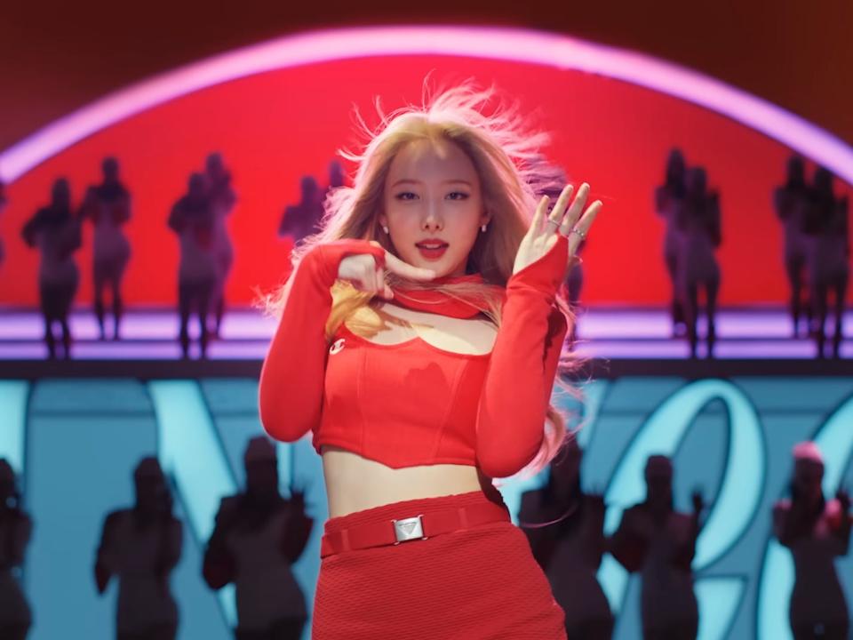 a young woman in a red outfit, standing in front of a two toned back ground filled with background dancers. she's making a gesture with her hands, with one pointing at the other, which is open